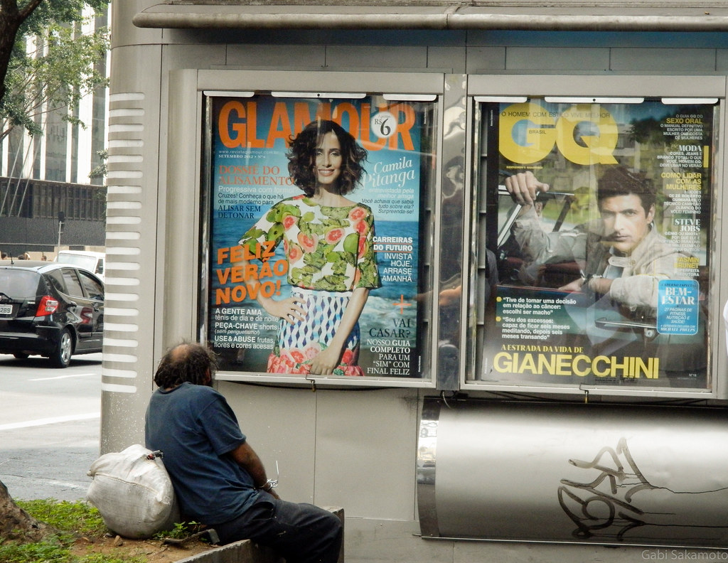 Um morador de rua sentado olhando os grandes pôsteres de revistas de cultura em uma banca de jornal