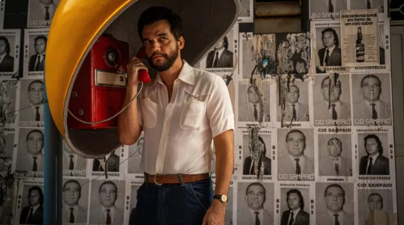 Wagner Moura de blusa branca em um orelhão, estampando o poster de O Agente Secreto