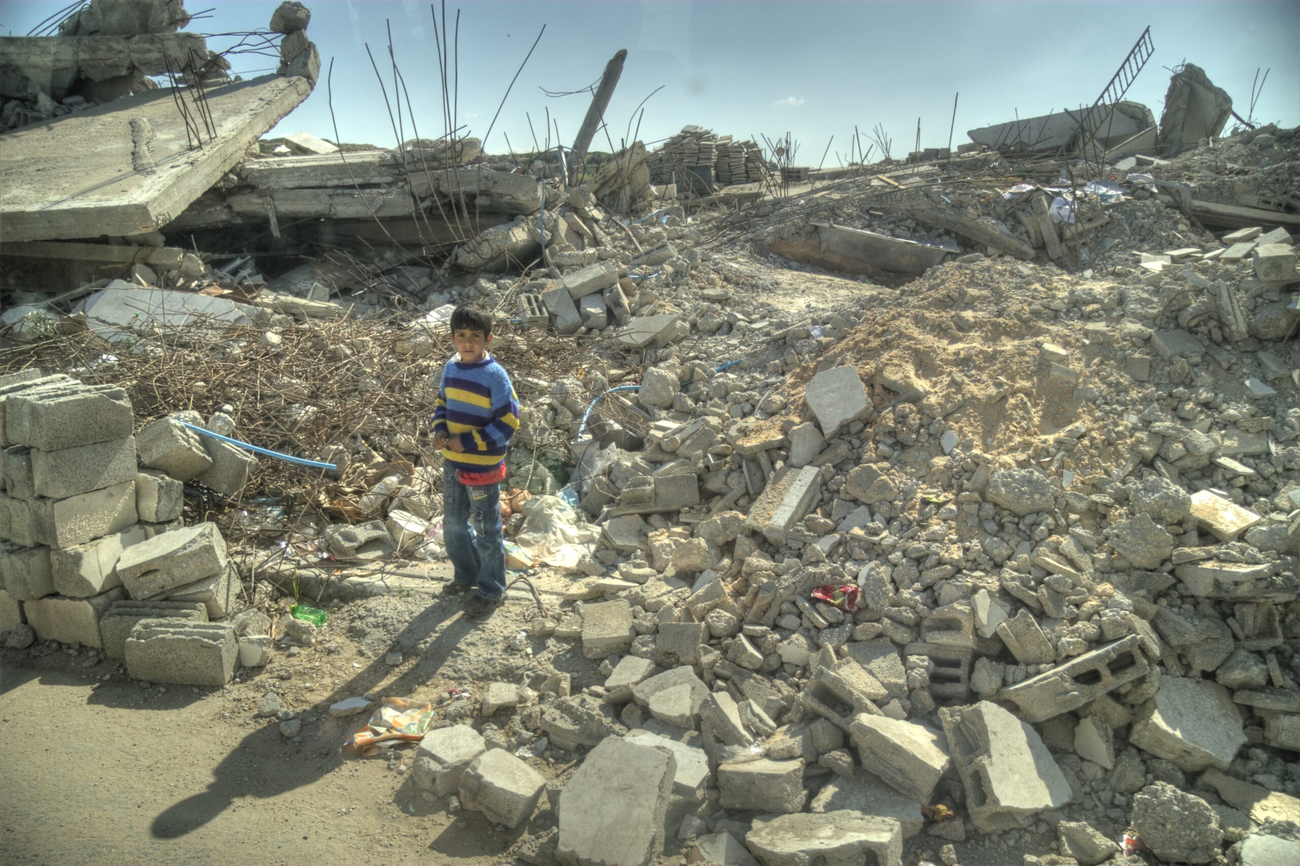 Cenário de Gaza: uma criança olha para a câmera em cima de destruição. A criança usa uma camiseta colorida.