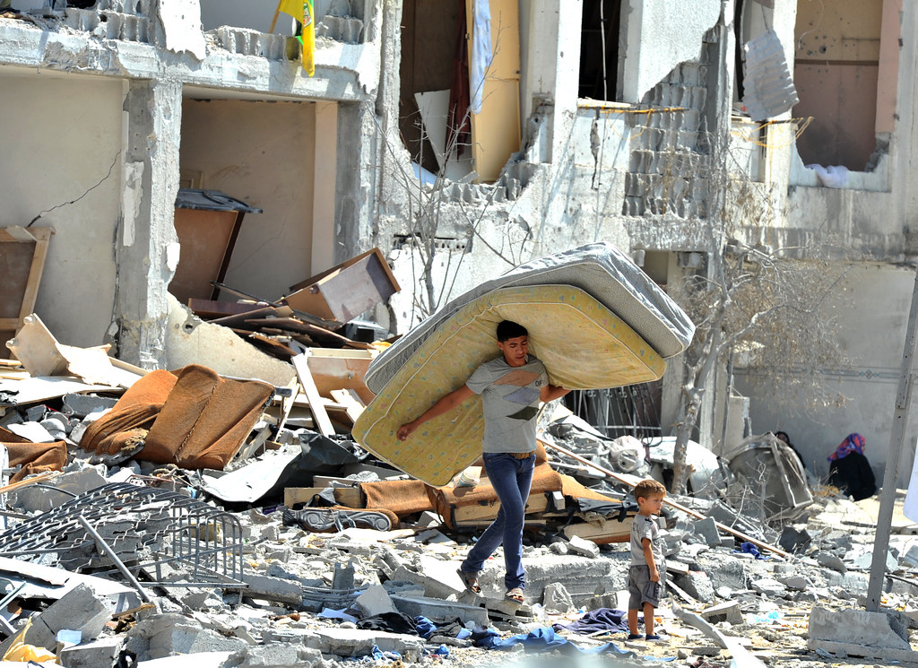 Genocídio em Gaza é ensaio militar. Homem carrega colchão com uma criança em meio à destruição.