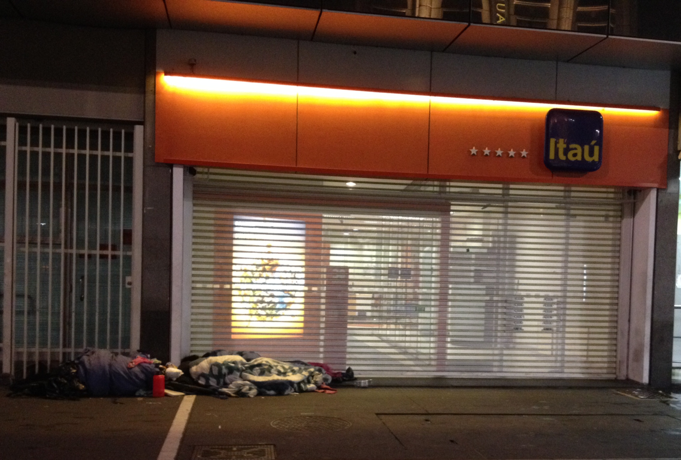 Imagem que representa a desigualdade do dinheiro: um morador de rua deitado em frente ao banco Itaú