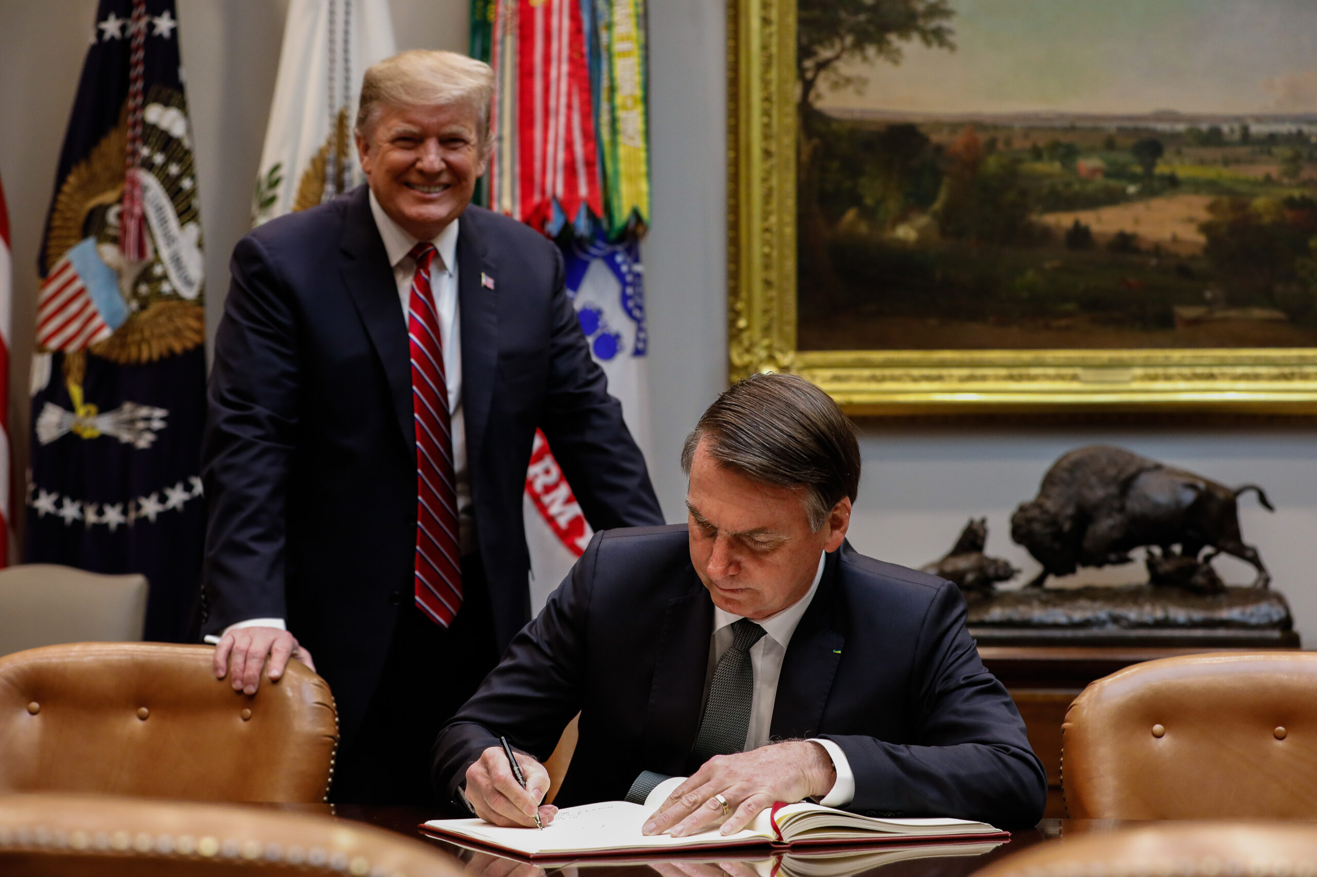 Trump sorri para a câmera enquanto Bolsonaro assina algo olhando para o papel.
