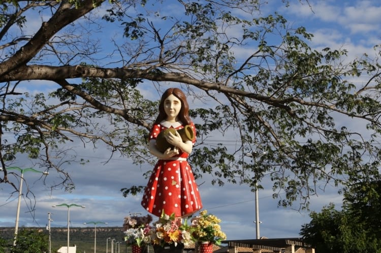 Estátua de Benigna Cardoso no Ceará abaixo de uma árvore. A menina usa um vestido vermelho com bolinhas brancas.