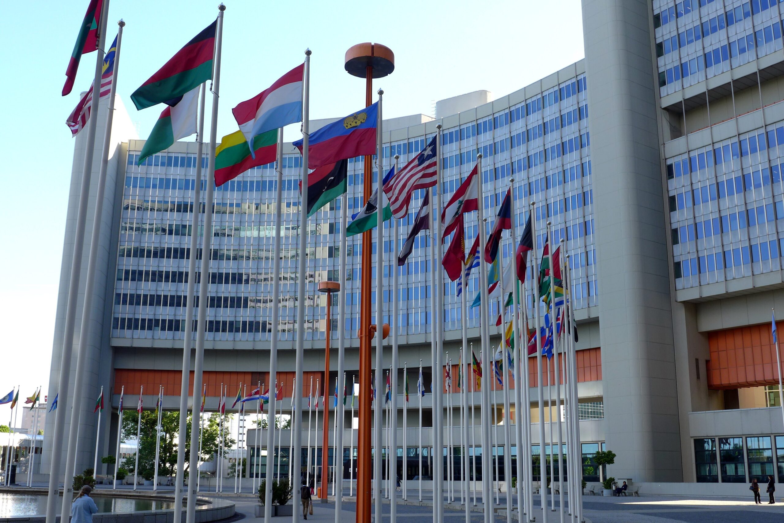 Parte da frente de uma das sedes da ONU. Na frente de um prédio, existem vários mastros com bandeiras.