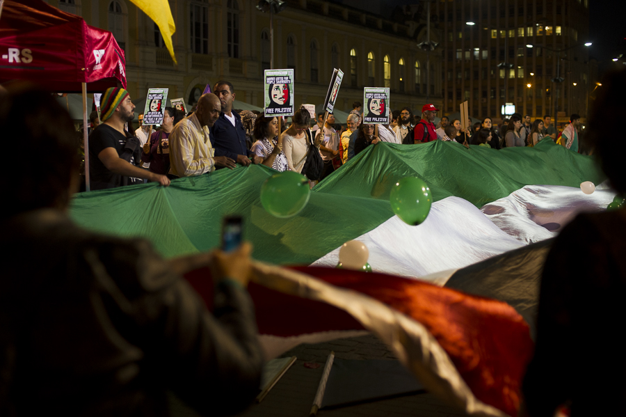 Manifestantes seguram uma bandeira palestina, enquanto levantam cartazem em que está escrita a frase "Free Palestine"
