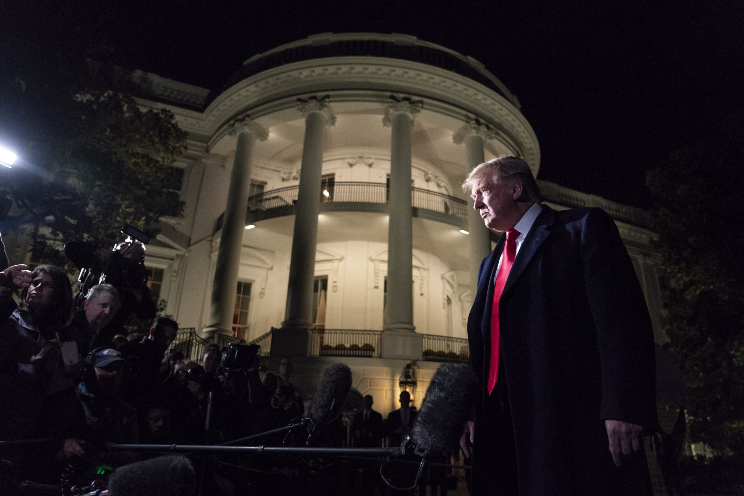 Imagem de Trump do lado de fora da Casa Branca à noite, cercado de fotógrafos.