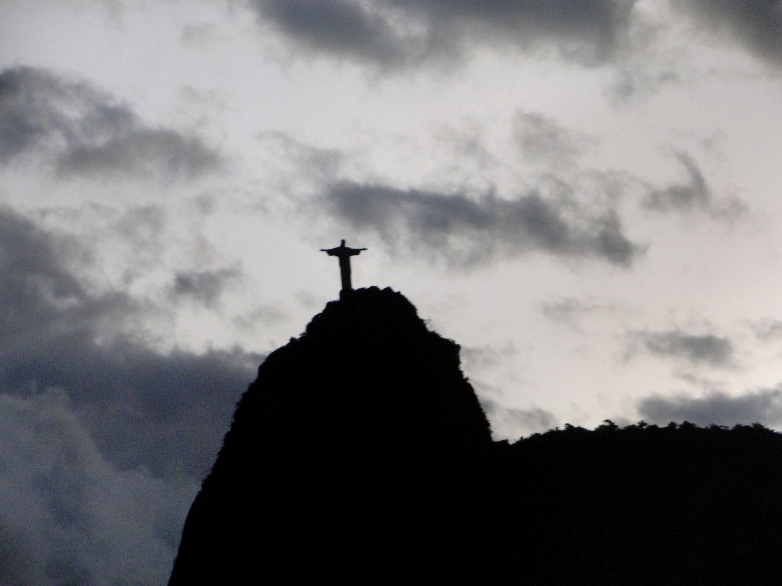 Fotografia do Cristo Redentor, no Rio de Janeiro. O céu está nublado, com muitas nuvens e o morro e o cristo sombreados.