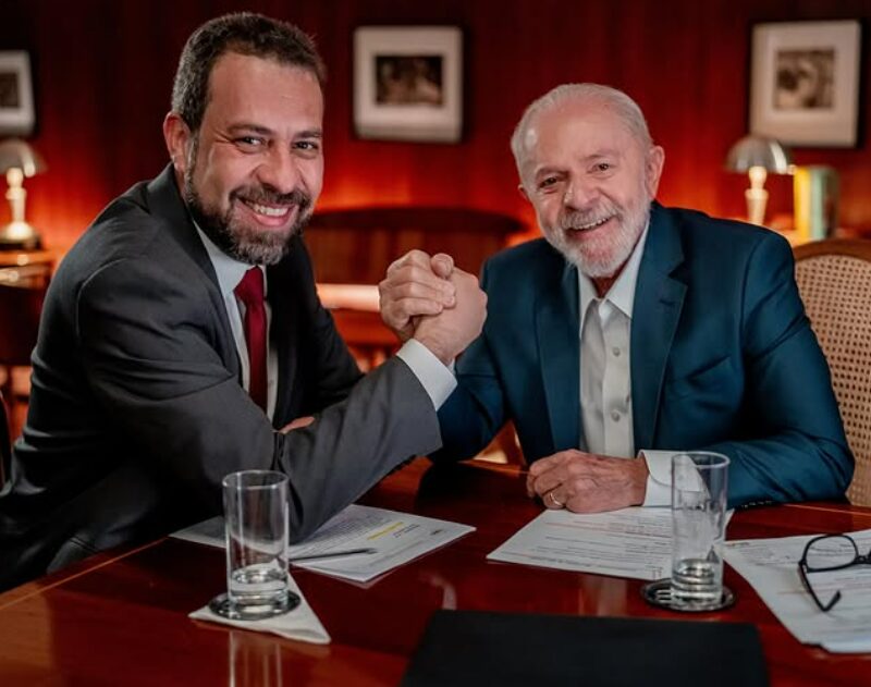Fotografia de Guilherme Boulos e Lula apertando as mãos. Ambos estão sentados e sorrindo.