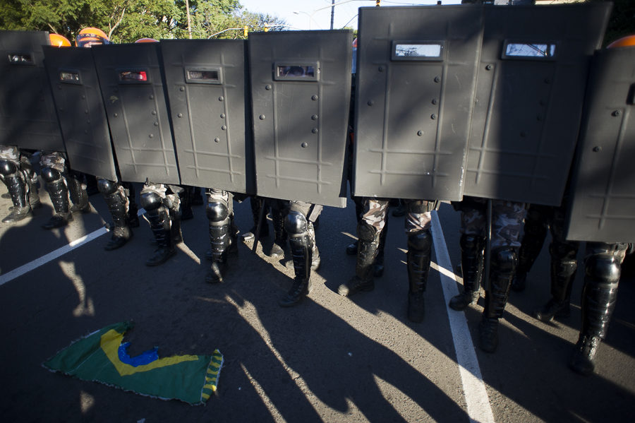 A economia política do massacre policial. A foto mostra vários escudos policiais em frente a uma bandeira.