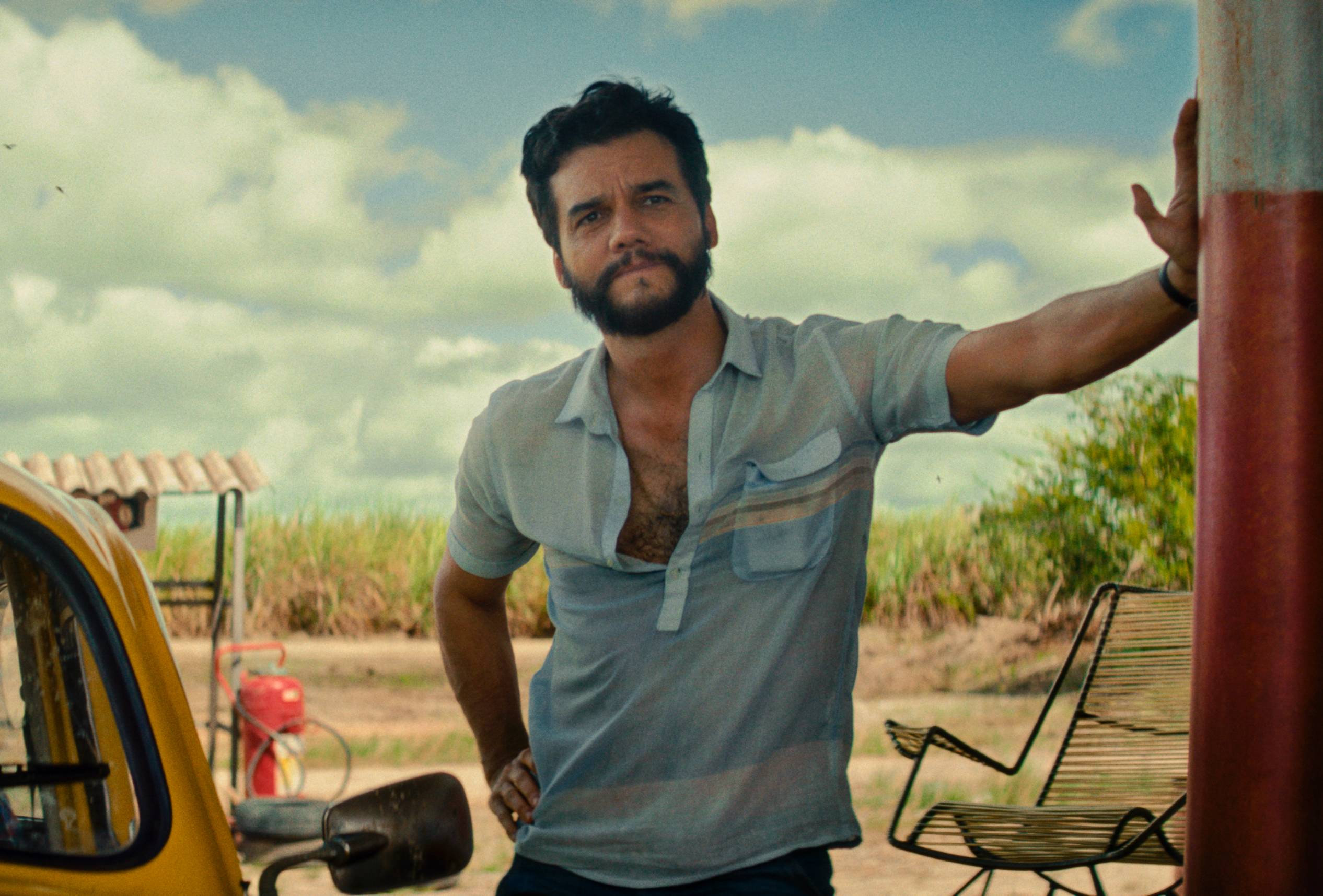 Fotografia de divulgação de O Agente Secreto. Wagner Moura usa uma blusa azul e se posiciona ao lado de um carro