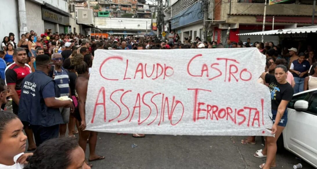 Manifestantes protestam a favor das vítimas. Levantam um cartaz escrito "Claudio Castro assassino terrorista"