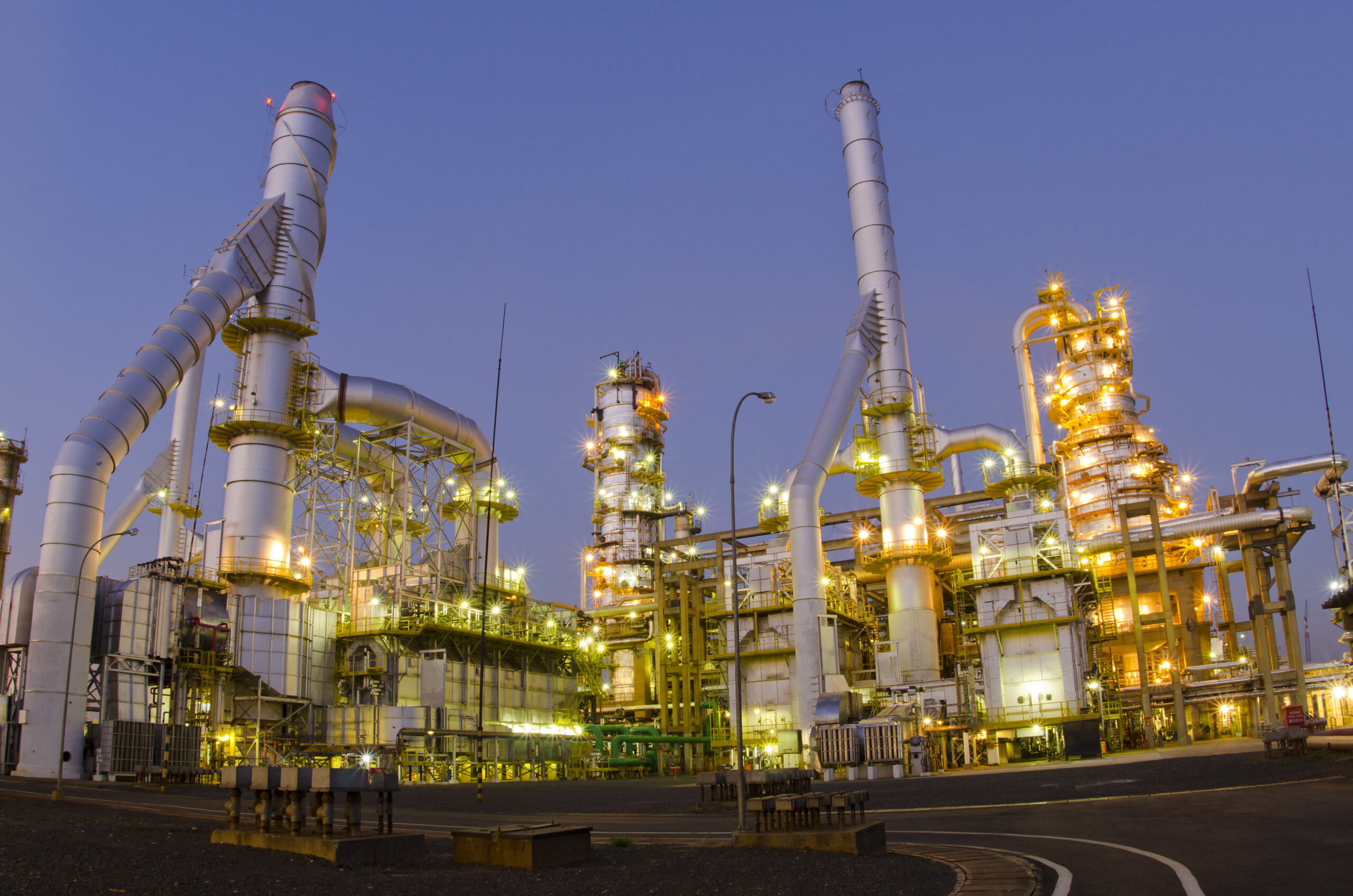 Refinaria de combustíveis. Existem várias luzes e o céu está escurecendo. Existem alguns carrinhos