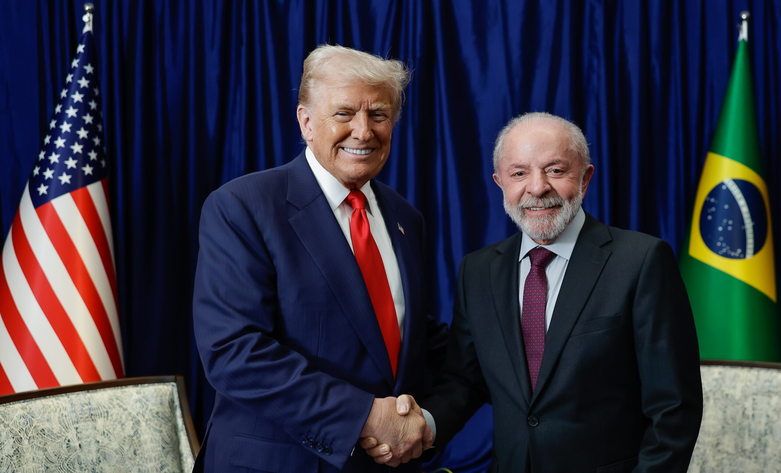 Trump e Lula segurando as mãos um do outros. Ambos sorriem. Do lado de cada um, estão as bandeiras de seus países.
