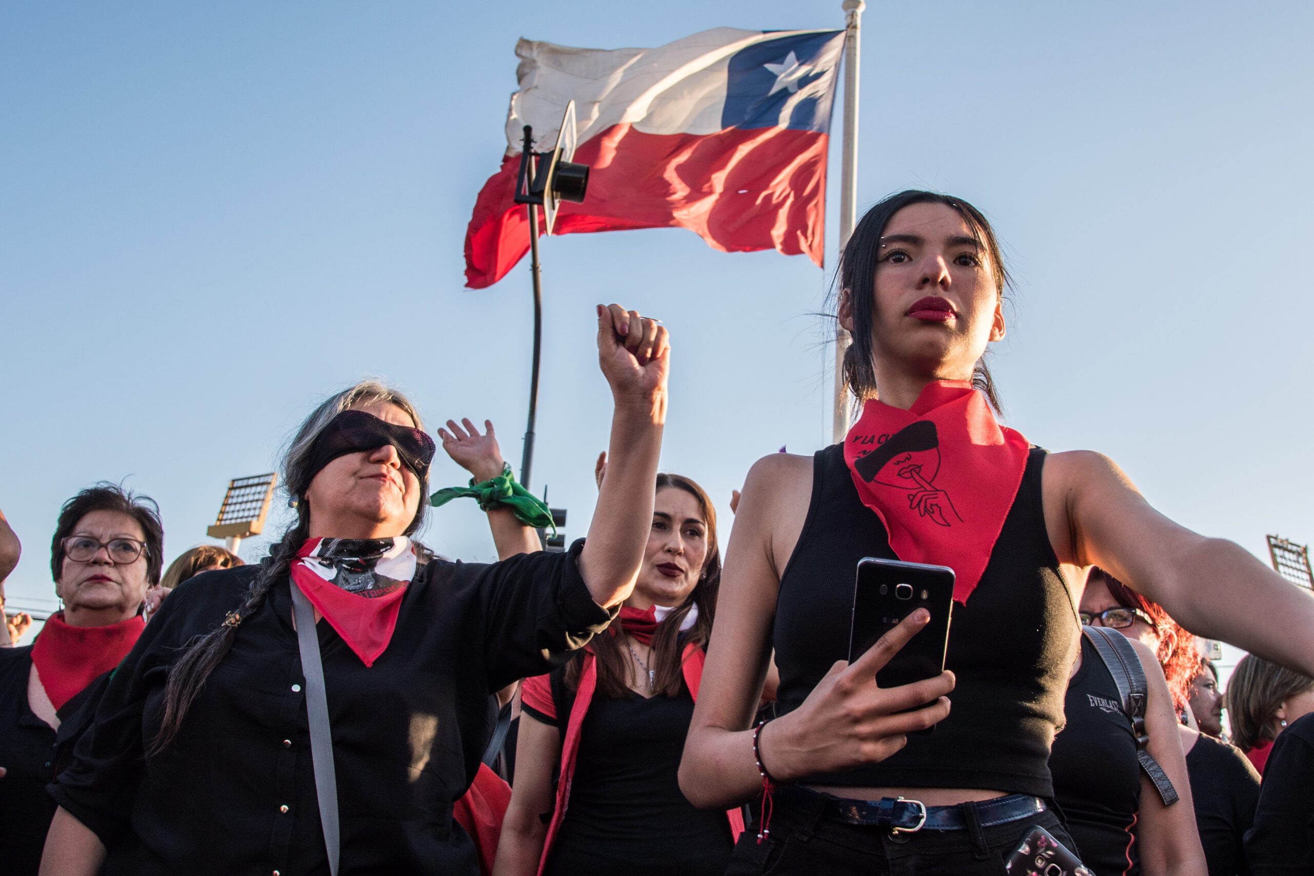 Mulheres protestam no Chile. Atrás delas, a bandeira chilena em um céu azul. Elas vestem preto e vermelho.