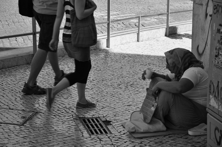 Entre sombras e permanências: quando a pobreza cai, mas permanece: fotografia de duas pessoas andando em frente a outra pessoa em situação de rua
