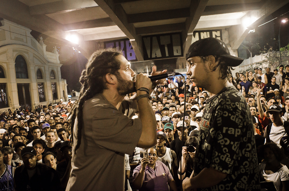 Dois MC´s em uma batalha de hip-hop. Eles estão em frente a uma grande plateia. Um está com o microfone, enquanto o outro aguarda olhando.