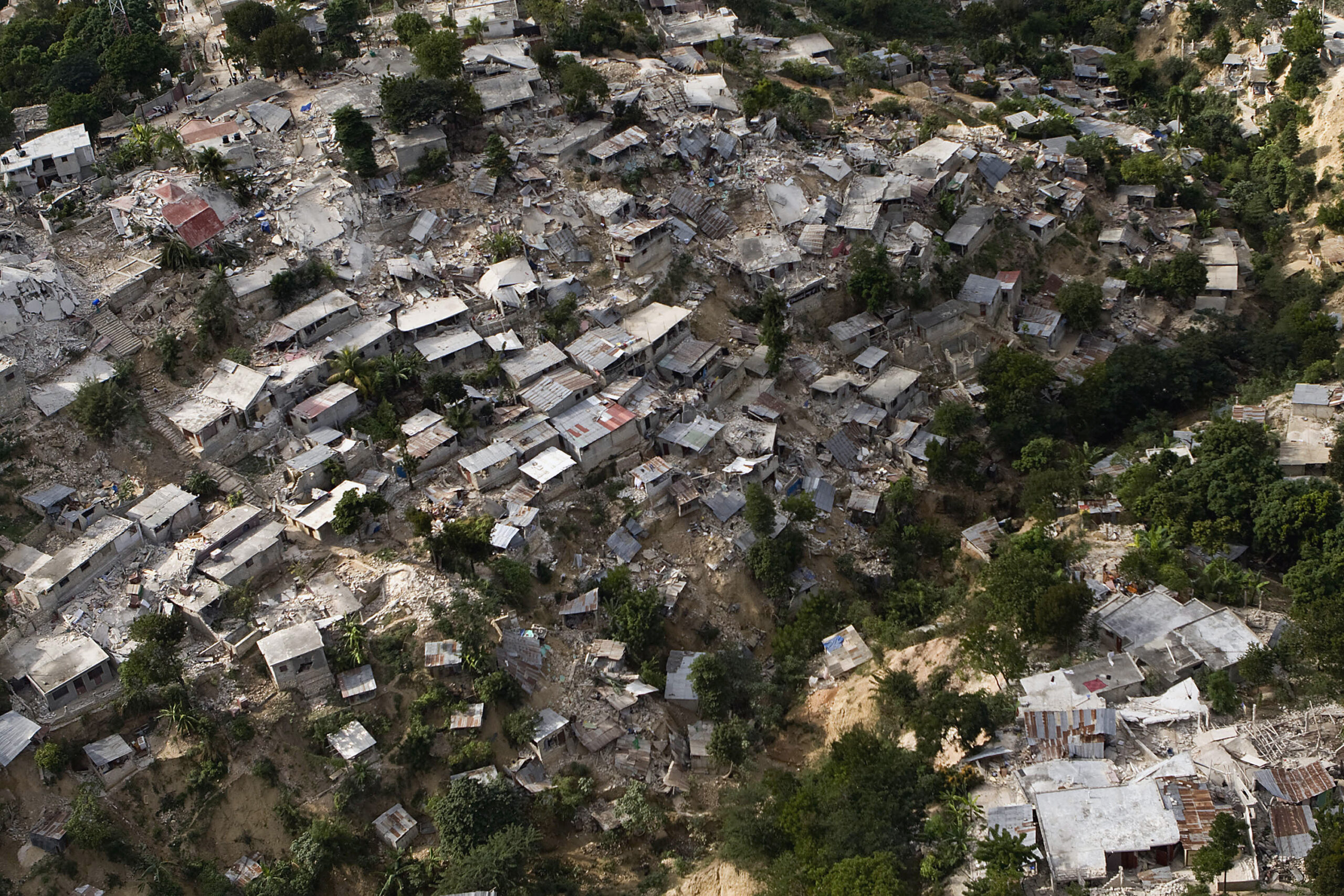 Fotografia de uma região haitiana após o terremoto de 2010. As casas e árvores estão derrubadas