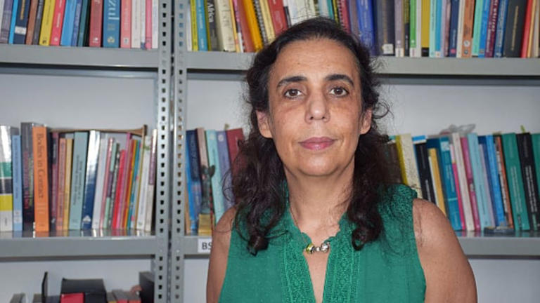 Fotografia de Maria Paula Meneses. Ela veste uma blusa verde e está na frente de uma prateleira