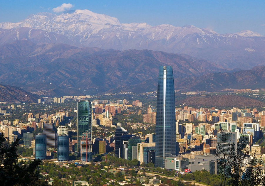 Fotografia da paisagem de Santiago, as cordilheiras estão ao fundo e há alguns prédios, sendo um deles o mais alto de todos