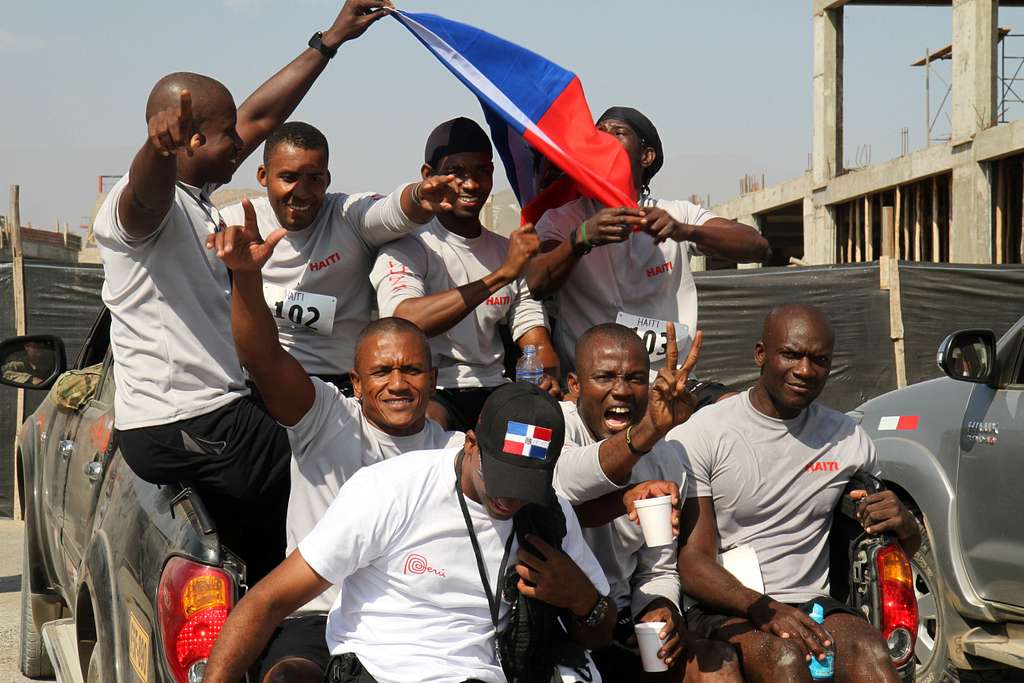 8 homens no Haiti estão na traseira de um carro, segurando a bandeira haitiana e sorrindo. Na blusa, escrito "Haiti"